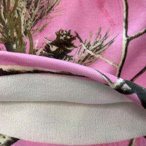 May include: A pink camouflage beanie hat with a white interior lining. The hat features a pink background with a pattern of brown and green tree branches. The hat is made of a soft, knitted material.