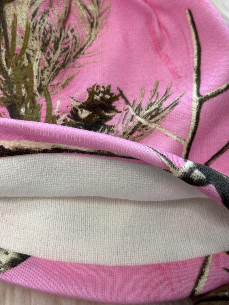 May include: A pink camouflage beanie hat with a white interior lining. The hat features a pink background with a pattern of brown and green tree branches. The hat is made of a soft, knitted material.