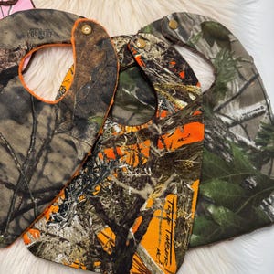 May include: Camouflage baby bibs in shades of brown, green, and orange. The bibs have snap closures and are made of a durable fabric. The bibs have a curved shape to catch spills and are designed for babies and toddlers.