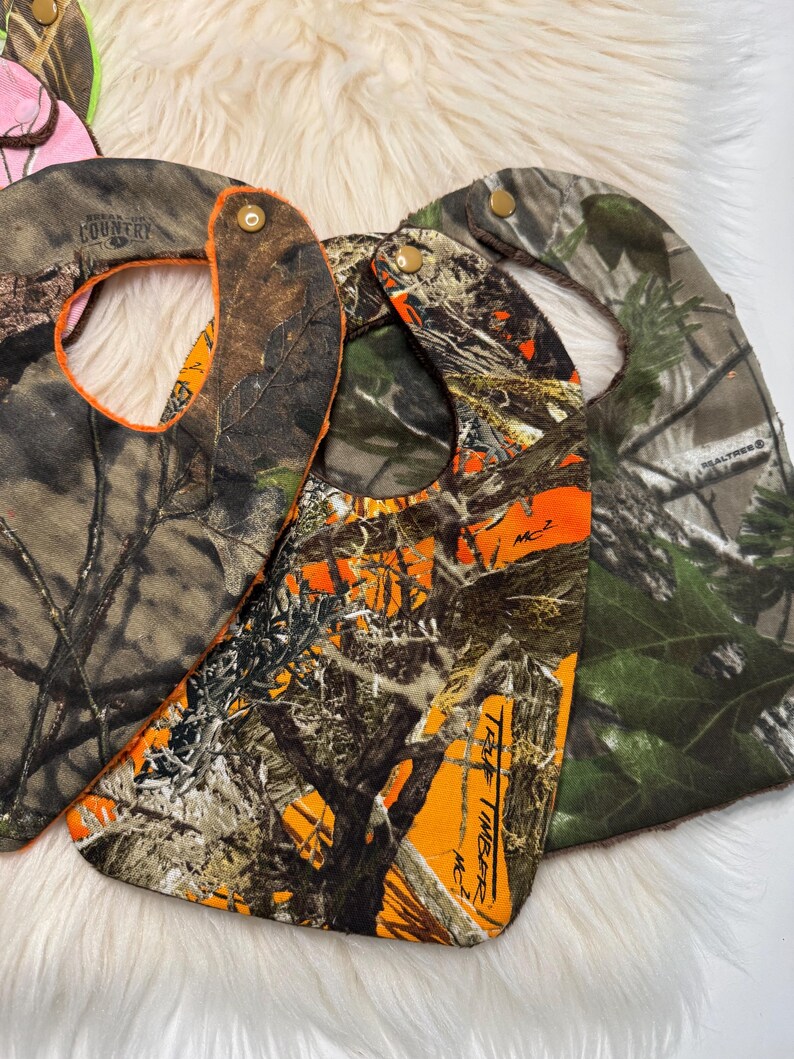 May include: Camouflage baby bibs in shades of brown, green, and orange. The bibs have snap closures and are made of a durable fabric. The bibs have a curved shape to catch spills and are designed for babies and toddlers.