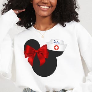 May include: White sweatshirt with a black and red Mickey Mouse head design. The Mickey Mouse head has a red bow and a white nurse's hat with a red cross and the name "Lisa" written on it.
