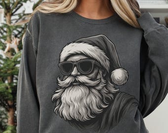 Pode incluir: Camisola cinza escuro com um gráfico a preto e branco do Pai Natal a usar óculos de sol e um gorro de Pai Natal. A ilustração do Pai Natal é um desenho detalhado. A camisola é combinada com calças de ganga azuis. Uma árvore de Natal está no fundo.