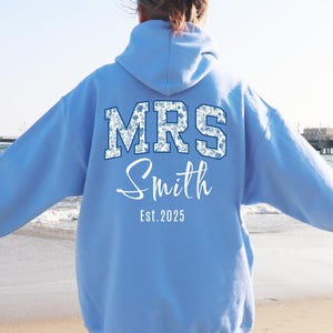 May include: A light blue hoodie with the text "MRS Smith" in a floral design on the back, with "Est. 2025" below. The hoodie is a solid color with a hood and long sleeves. The background shows a beach and ocean.
