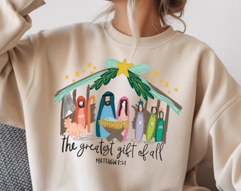 Pode incluir: Camisola bege com uma ilustração colorida de uma cena de presépio. O design inclui o texto "the greatest gift of all" e "Matthew 1:21". O gráfico mostra um estábulo, figuras e uma estrela.