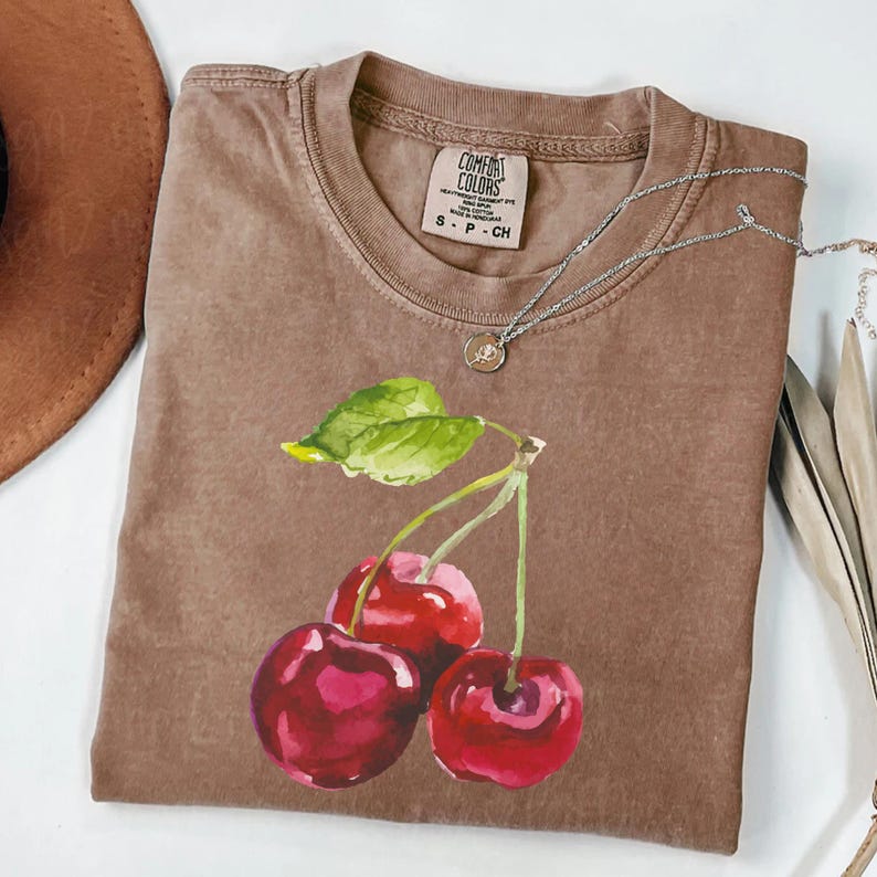May include: A brown t-shirt featuring a watercolour illustration of three red cherries and green leaves. The shirt has the text "COMFORT COLORS" and "S - P - CH" printed on the tag.