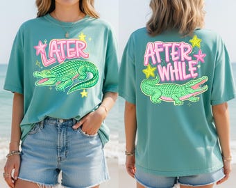 Pode incluir: Camiseta verde-azulado com um gráfico de jacaré verde e a palavra "LATER" em rosa na frente. A parte de trás apresenta as palavras "AFTER WHILE" e um design de jacaré correspondente. A camiseta tem um corte folgado.
