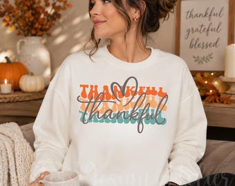 Könnte beinhalten: Ein cremefarbenes Sweatshirt mit Rundhalsausschnitt und einem Grafikdruck, bei dem das Wort Thankful in wellenförmigen horizontalen Bändern in Orange, Gelb und Blaugrün wiederholt wird, ergänzt durch ein dunkelgraues Herz.