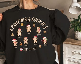 Pode incluir: Camisola preta com o texto "Grandma's Cookies" num design circular. Ilustrações de biscoitos de gengibre com nomes são apresentadas abaixo do texto. A camisola também inclui o texto "Est. 2025".
