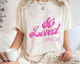 Pode incluir: Camiseta creme com as palavras "So Loved" em rosa, acentuada por pequenos padrões florais rosa. O texto "John 3:16" está abaixo. A camiseta é de estilo casual.