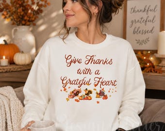 Könnte beinhalten: Ein weißes Sweatshirt mit Rundhalsausschnitt und dem braunen Schriftzug Give Thanks with a Grateful Heart. Das Design enthält bunte Illustrationen von Truthähnen, Kürbissen und Herbstblättern auf der Brust. Das Kleidungsstück hat lange Ärmel.