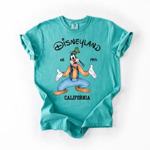 Peut inclure: T-shirt turquoise avec un dessin de Dingo de Disney, et le texte "Disneyland est 1955 California". Dingo porte une chemise orange, un pantalon bleu et un chapeau vert. Le t-shirt a des manches retrouss&eacute;es.