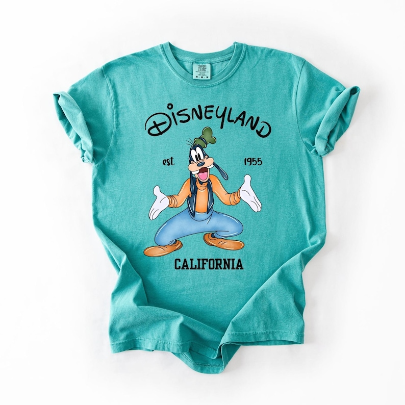 Peut inclure: T-shirt turquoise avec un dessin de Dingo de Disney, et le texte "Disneyland est 1955 California". Dingo porte une chemise orange, un pantalon bleu et un chapeau vert. Le t-shirt a des manches retrouss&eacute;es.