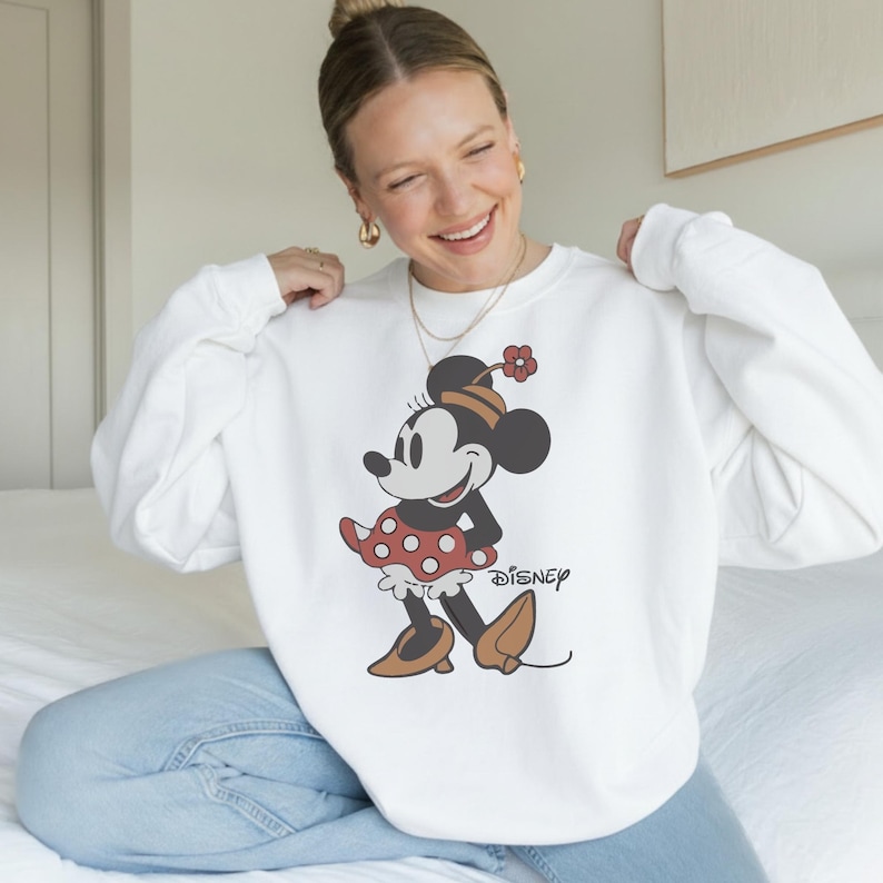 May include: White sweatshirt featuring a graphic of Minnie Mouse in a red polka dot dress, brown shoes, and a hat with a flower. The word "Disney" is printed below the character.