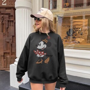 May include: Black sweatshirt featuring a Minnie Mouse graphic. The graphic shows Minnie Mouse in a red polka dot dress, brown shoes, and a hat with a flower. The word "Disney" is printed below the graphic.