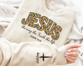 May include: Beige sweatshirt with the word "JESUS" in leopard print, with the text "the way the Truth the life" and "JOHN 14:6". The sleeve has a cross graphic and the name "Ashley".