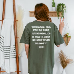 Könnte beinhalten: Olivgrünes T-Shirt mit weißem Text: "WE MUST ESPECIALLY BEWARE OF THAT SMALL GROUP OF SELFISH MEN WHO WOULD CLIP THE WINGS OF THE AMERICAN EAGLE IN ORDER TO FEATHER THEIR OWN NESTS." -FDR 1941. Das Shirt hat eine lockere Passform.