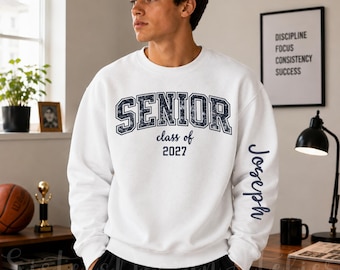 Pode incluir: Uma camisola branca de gola redonda com a palavra SENIOR numa fonte azul escura desgastada no peito, com o texto class of 2027 por baixo. O nome Joseph está impresso numa fonte cursiva azul escura na manga esquerda.