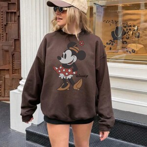 May include: A brown sweatshirt featuring a Minnie Mouse graphic. The cartoon character is depicted in a red and white polka dot dress, with a flower on her head. The word "Disney" is printed below the character.