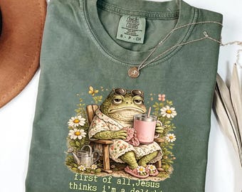 Pode incluir: Uma t-shirt verde-oliva com o gráfico de um sapo usando óculos de sol e um roupão floral, sentado numa cadeira de madeira enquanto segura uma caneca rosa. O texto abaixo diz, first of all, Jesus thinks i'm a delight. Gola redonda.