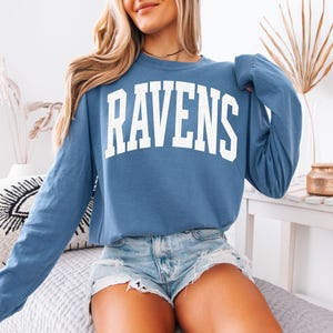 Puede incluir: Una sudadera azul de manga larga con la palabra "RAVENS" en letras blancas grandes en el pecho. La sudadera es de color azul liso y tiene cuello redondo. La modelo lleva pantalones cortos vaqueros.