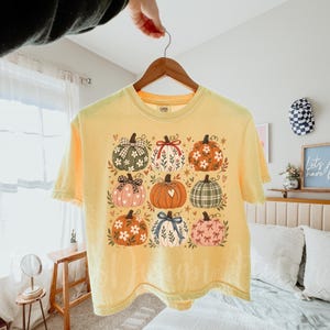 Pode incluir: Uma t-shirt amarelo-pálido com uma impressão gráfica de nove abóboras estilizadas dispostas numa grelha. Cada abóbora apresenta padrões únicos, incluindo motivos florais, xadrez, bolinhas e laços em tons de laranja, verde, rosa e branco.