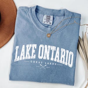 May include: A blue short-sleeved t-shirt with a distressed white graphic that reads LAKE ONTARIO GREAT LAKES EST. 1815 with a crossed oar icon. The shirt features a Comfort Colors brand label at the inner neckline and a small gold-toned pendant necklace.