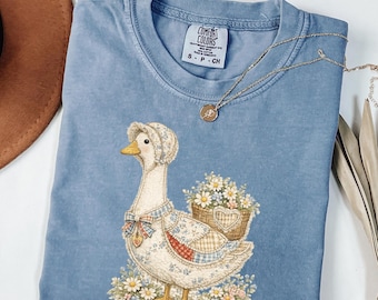 Pode incluir: Camiseta azul Comfort Colors com uma ilustração de um ganso usando um chapéu e carregando uma cesta de flores. O ganso é adornado com um design de patchwork. Um colar de ouro com um pingente repousa na camisa.