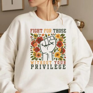 May include: Cream-colored sweatshirt with the text "FIGHT FOR THOSE WITHOUT YOUR PRIVILEGE" in a bold, multi-colored font. The design features a raised fist surrounded by red, orange, and yellow flowers and green leaves.