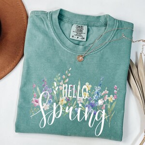 Puede incluir: Una camiseta Comfort Colors verde azulado con un diseño floral y las palabras "Hello Spring" en escritura blanca. Un collar plateado con un pequeño colgante descansa sobre la camisa. La camisa es talla S.