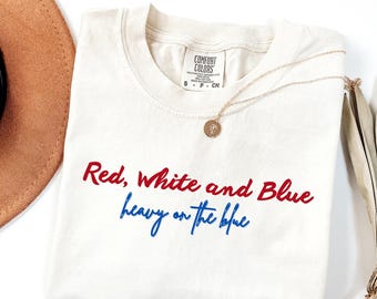 May include: Cream-colored t-shirt with the text "Red, White and Blue heavy on the blue" in red and blue script. A gold necklace with a pendant and a brown hat are also in the image.