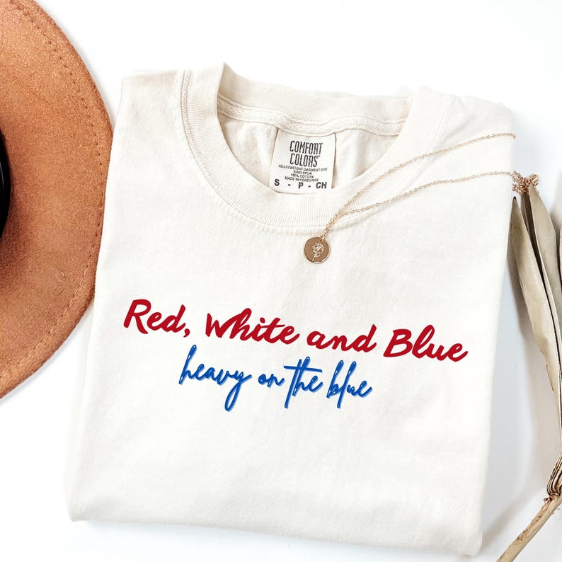 May include: Cream-colored t-shirt with the text "Red, White and Blue heavy on the blue" in red and blue script. A gold necklace with a pendant and a brown hat are also in the image.