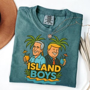 Puede incluir: Camiseta Comfort Colors de color verde azulado con un dibujo animado de dos hombres sosteniendo bebidas tropicales bajo palmeras. El texto "ISLAND BOYS" está en amarillo y azul. Un collar plateado con un colgante está sobre la camiseta.