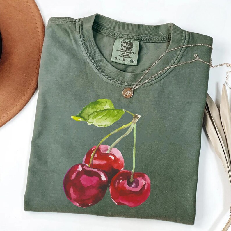 May include: A green t-shirt featuring a watercolour painting of three red cherries and green leaves. The shirt has a tag that reads "Comfort Colors" and "S-P-CH".