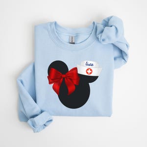 May include: Light blue sweatshirt with a black and red Mickey Mouse design. The Mickey Mouse has a red bow and a white nurse's hat with a red cross and the name "Lisa" on it.