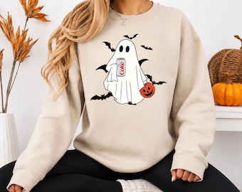 Op de afbeelding: Een beige sweater met ronde hals voorzien van een grafische print van een witte geest die een blikje Diet Coke en een oranje pompoen vasthoudt. Het ontwerp bevat diverse zwarte silhouetten van vliegende vleermuizen rondom de geest.