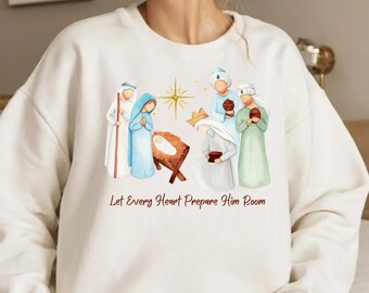 Pode incluir: Camisola creme com uma ilustração em aquarela da cena da Natividade, com Maria, José e o menino Jesus. O design inclui uma estrela e o texto "Let Every Heart Prepare Him Room."
