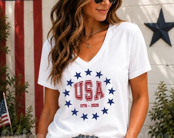 Pode incluir: Uma t-shirt branca de manga curta com decote em V, apresentando um gráfico circular de estrelas azuis em torno do texto vermelho USA e das datas 1776-2026. A t-shirt é feita de um tecido leve e tem um corte clássico e simples.