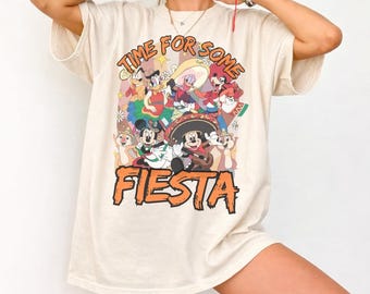 Pode incluir: Camiseta creme com um gráfico colorido de personagens de desenhos animados em trajes festivos. O texto "TIME FOR SOME FIESTA" está arqueado acima dos personagens, com "FIESTA" em letras grandes laranja abaixo.