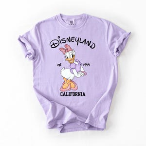 Peut inclure: Un t-shirt violet clair avec Daisy Duck de Disneyland. Le t-shirt porte le logo Disneyland, le texte "est. 1955" et "CALIFORNIA" imprim&eacute; en noir. Daisy Duck porte un n&oelig;ud et une robe roses.