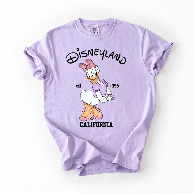 Peut inclure: Un t-shirt violet clair avec Daisy Duck de Disneyland. Le t-shirt porte le logo Disneyland, le texte "est. 1955" et "CALIFORNIA" imprim&eacute; en noir. Daisy Duck porte un n&oelig;ud et une robe roses.