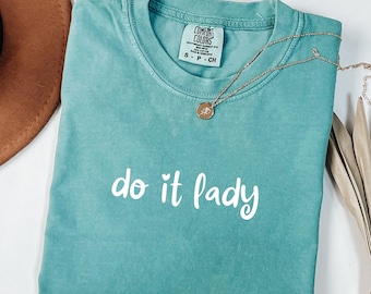 Puede incluir: Una camiseta turquesa Comfort Colors con la frase "do it lady" en escritura blanca. Un collar dorado con un colgante descansa sobre la camiseta. La camiseta está doblada y un sombrero marrón es parcialmente visible en el fondo.