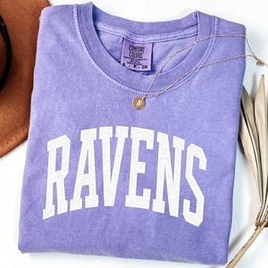 Puede incluir: Una camiseta lavanda Comfort Colors con la palabra "RAVENS" en letras blancas. Un collar de oro con un colgante descansa sobre la camiseta. La camiseta est&aacute; doblada cuidadosamente sobre una superficie blanca.