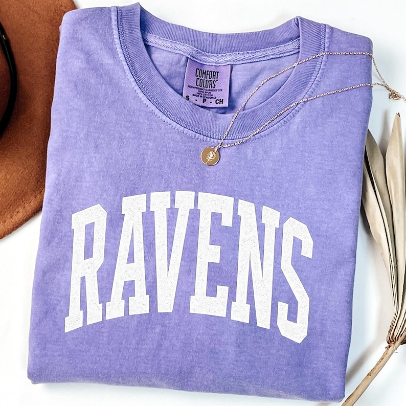 Puede incluir: Una camiseta lavanda Comfort Colors con la palabra "RAVENS" en letras blancas. Un collar de oro con un colgante descansa sobre la camiseta. La camiseta est&aacute; doblada cuidadosamente sobre una superficie blanca.