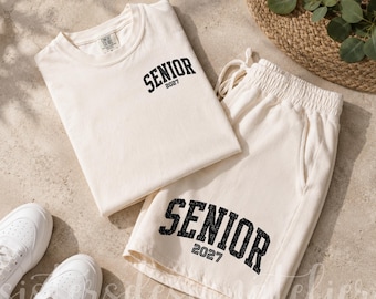 Pode incluir: Um conjunto de t-shirt e calções cor de creme com letras pretas desgastadas que dizem SENIOR 2027. A t-shirt tem uma pequena impressão no peito, enquanto os calções apresentam uma versão maior do mesmo texto na parte inferior da perna.