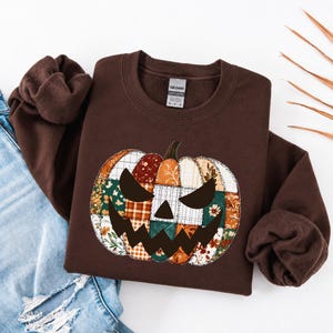 Op de afbeelding: Een donkerbruine sweater met ronde hals en een grote, centrale afbeelding van een pompoen gemaakt van verschillende patchwork-stofpatronen in tinten oranje, groen en wit, met een donkerbruin, grillig, uitgesneden pompoengezicht.