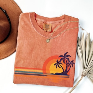 May include: A coral orange t-shirt with a vintage sunset graphic featuring palm trees and a beach scene. The shirt has a crew neck and is made of a soft, comfortable fabric. The text "COMFORT COLORS" is visible on the tag.