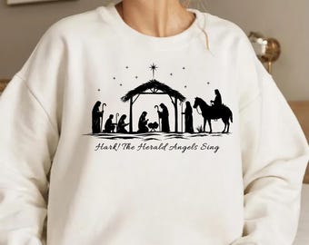 Pode incluir: Camisola creme com uma silhueta preta da cena da Natividade, incluindo Maria, José e o menino Jesus. O texto "Hark! The Herald Angels Sing" está impresso abaixo. Uma estrela está acima do estábulo.