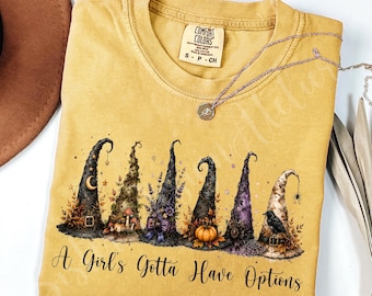 Pode incluir: Uma t-shirt amarelo-mostarda com um gráfico de seis chapéus de bruxa distintos decorados com elementos de outono como abóboras, folhas e um corvo. O texto "A Girl's Gotta Have Options" está impresso em letra preta sob os chapéus.
