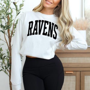 Puede incluir: Sudadera blanca de manga larga con la palabra "RAVENS" en letras negras en negrita en el pecho. Se combina con leggings negros. El estilo general es casual y deportivo.