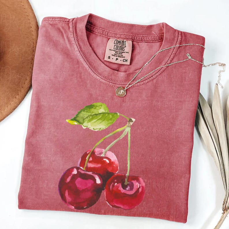 May include: A red t-shirt featuring a watercolour painting of three cherries and a green leaf. The shirt has the text "COMFORT COLORS" on the tag.
